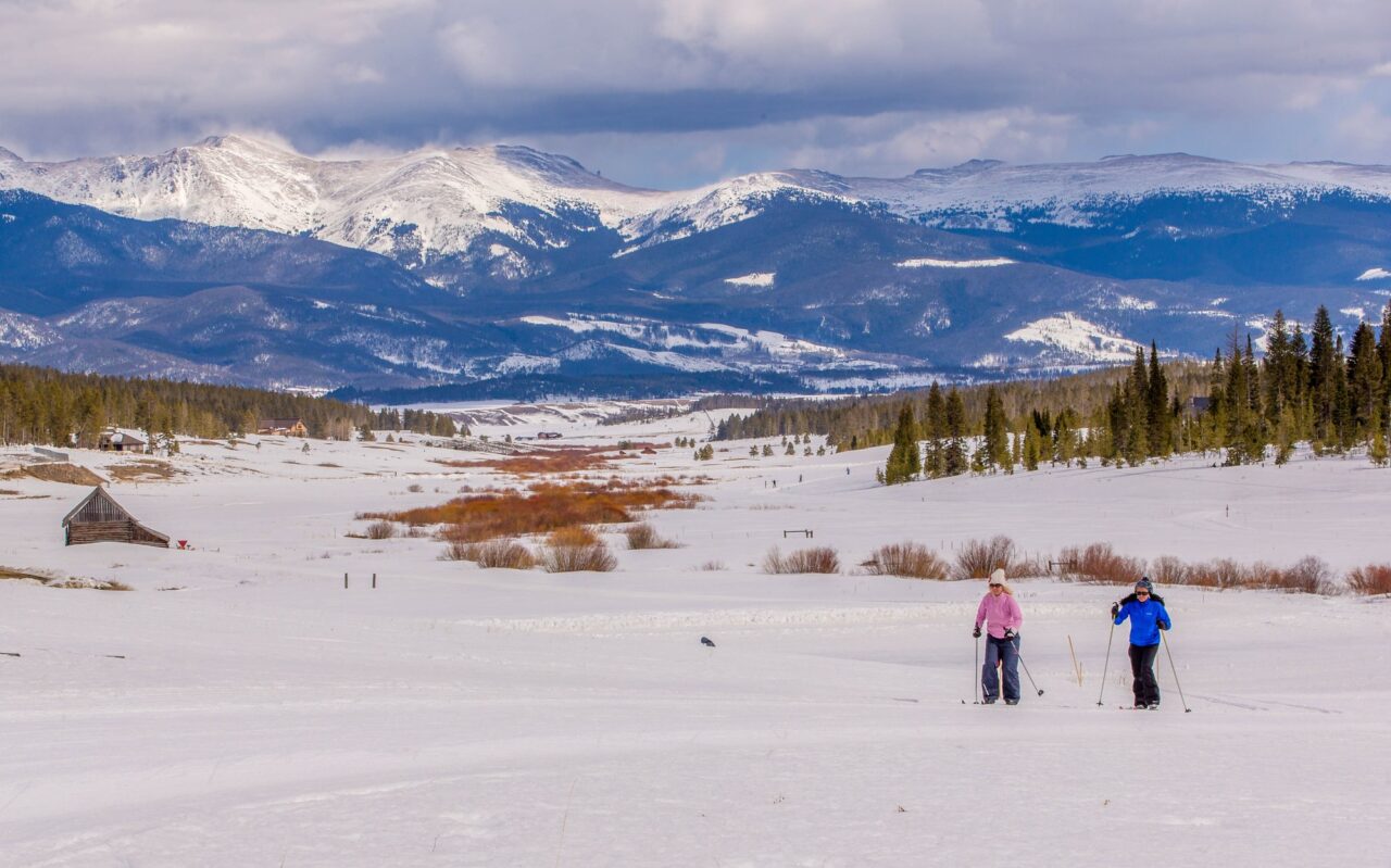 Two skiers cross a wide snow-covered meadow at Snow Mountain Ranch, with a snow-capped mountain range under cloudy skies behind them.