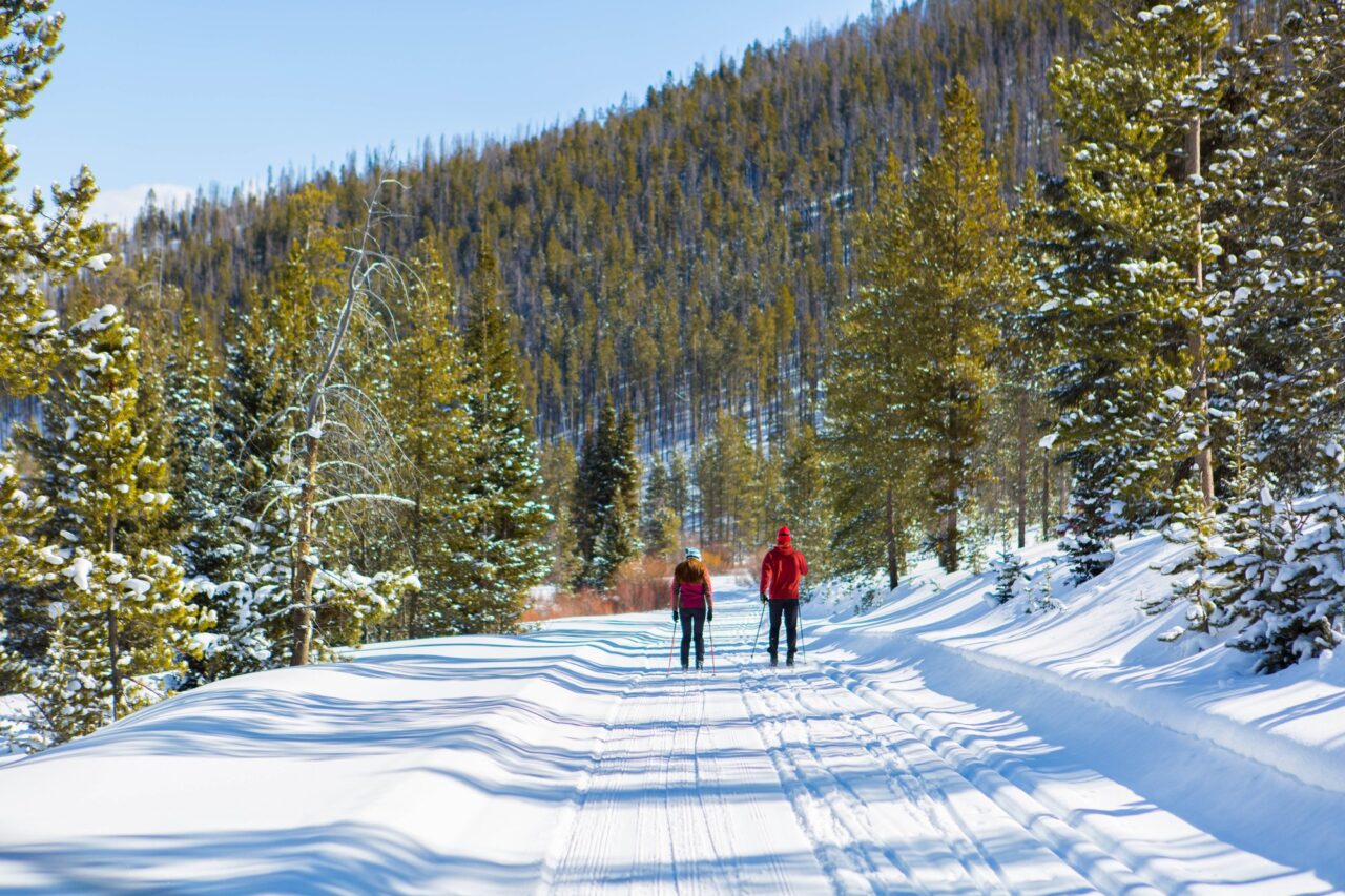 Two skiers on a groomed trail between snowy pines at Snow Mountain Ranch, one in a red jacket and one in a pink jacket.