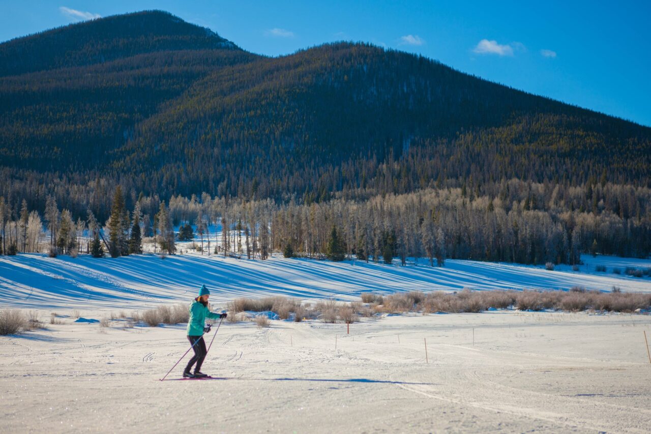 A lone skier in a teal jacket crosses an open snowfield below forested hills at Snow Mountain Ranch on a clear day.