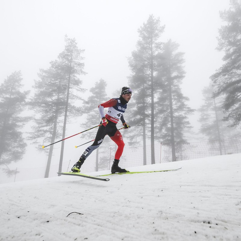 Thomas Stephen in a red and black Canada suit skating up a snowy slope with tall pines fading into fog