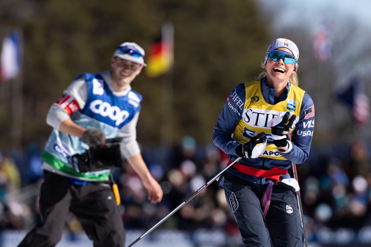 Jessie Diggins laughs on a warm-up lap while a filmmaker in a media bib skis alongside her carrying a camera, a blurred crowd behind them.