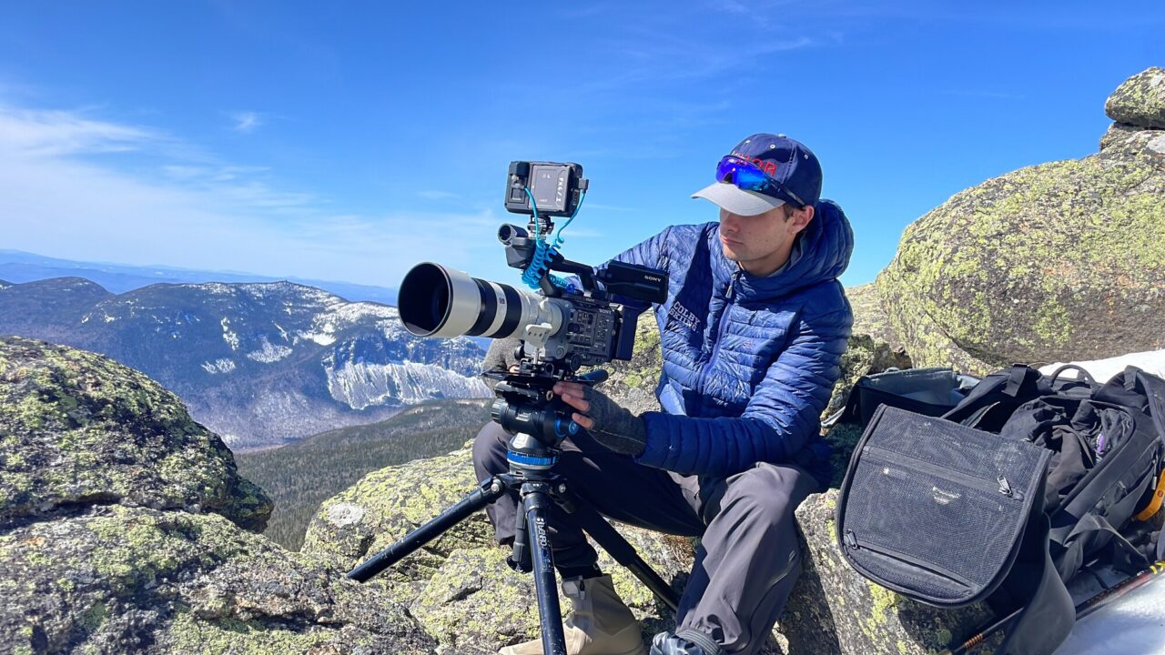 A filmmaker sits on a rocky summit behind a tripod-mounted cinema camera with a long telephoto lens, snow-streaked mountains behind him.