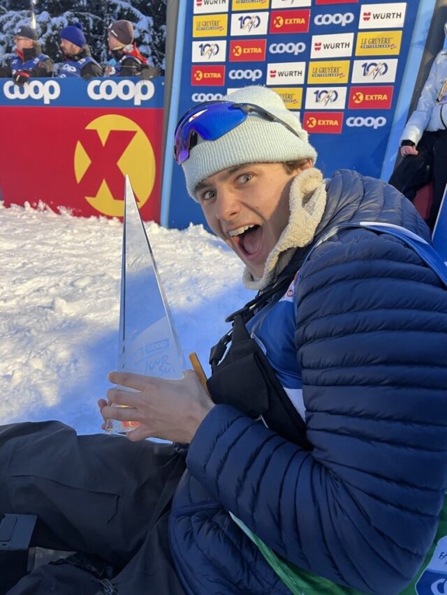 A filmmaker sitting in the snow holding a tall glass pyramid trophy, with race sponsor banners behind him.