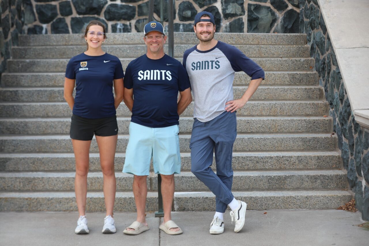 Three St. Scholastica coaches in Saints shirts standing at the foot of a wide stone staircase, Chad Salmela in the middle in a cap and light blue shorts.