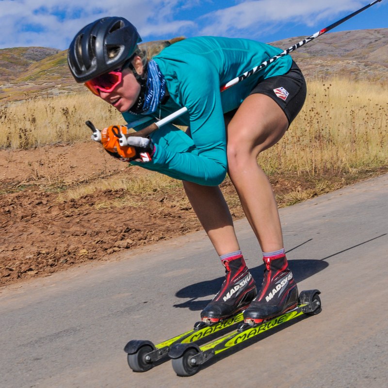 Hailey Swirbul in a teal top, black helmet and orange goggles crouched low on rollerskis on a paved road with dry grassy hills behind