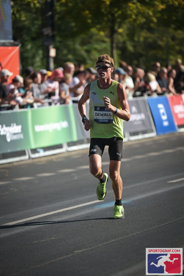 Lars Dewall in a lime green singlet and bright yellow shoes running alone down a sunny road lined with barriers and spectators.