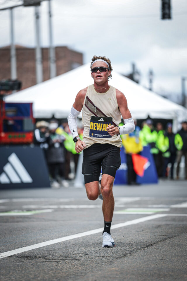 Lars Dewall in a cream singlet with a maroon sash, white arm sleeves, headband and dark sunglasses running toward the camera at the Boston Marathon, a white tent and race volunteers behind him.