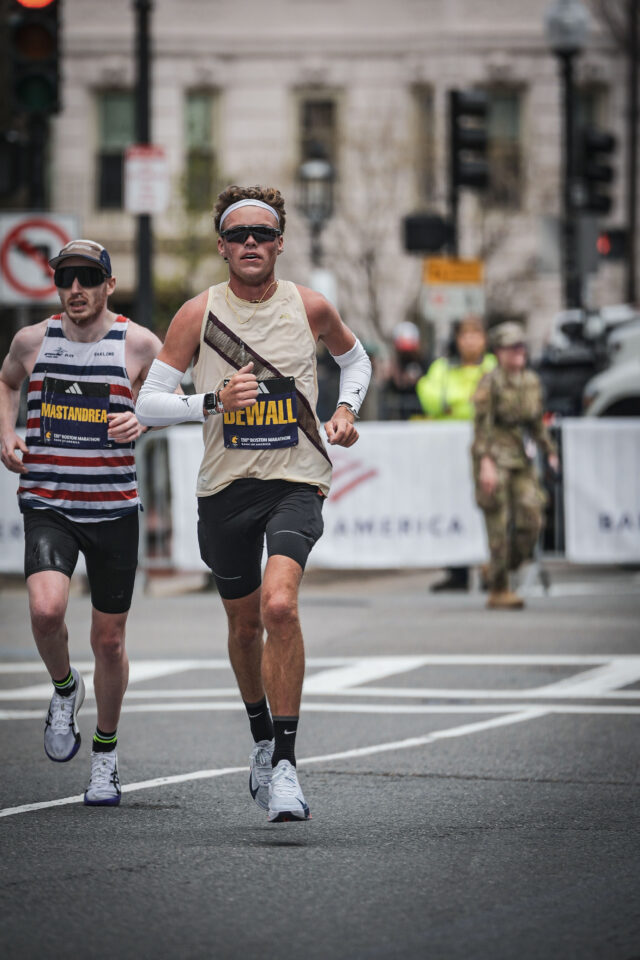 Lars Dewall in a cream singlet and white arm sleeves running a city street at the Boston Marathon, a runner in a striped singlet a step behind him.