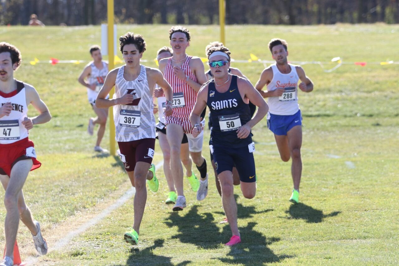 Lars Dewall in a navy St. Scholastica Saints singlet and bib 50 running in a pack of college runners on a grass cross country course.