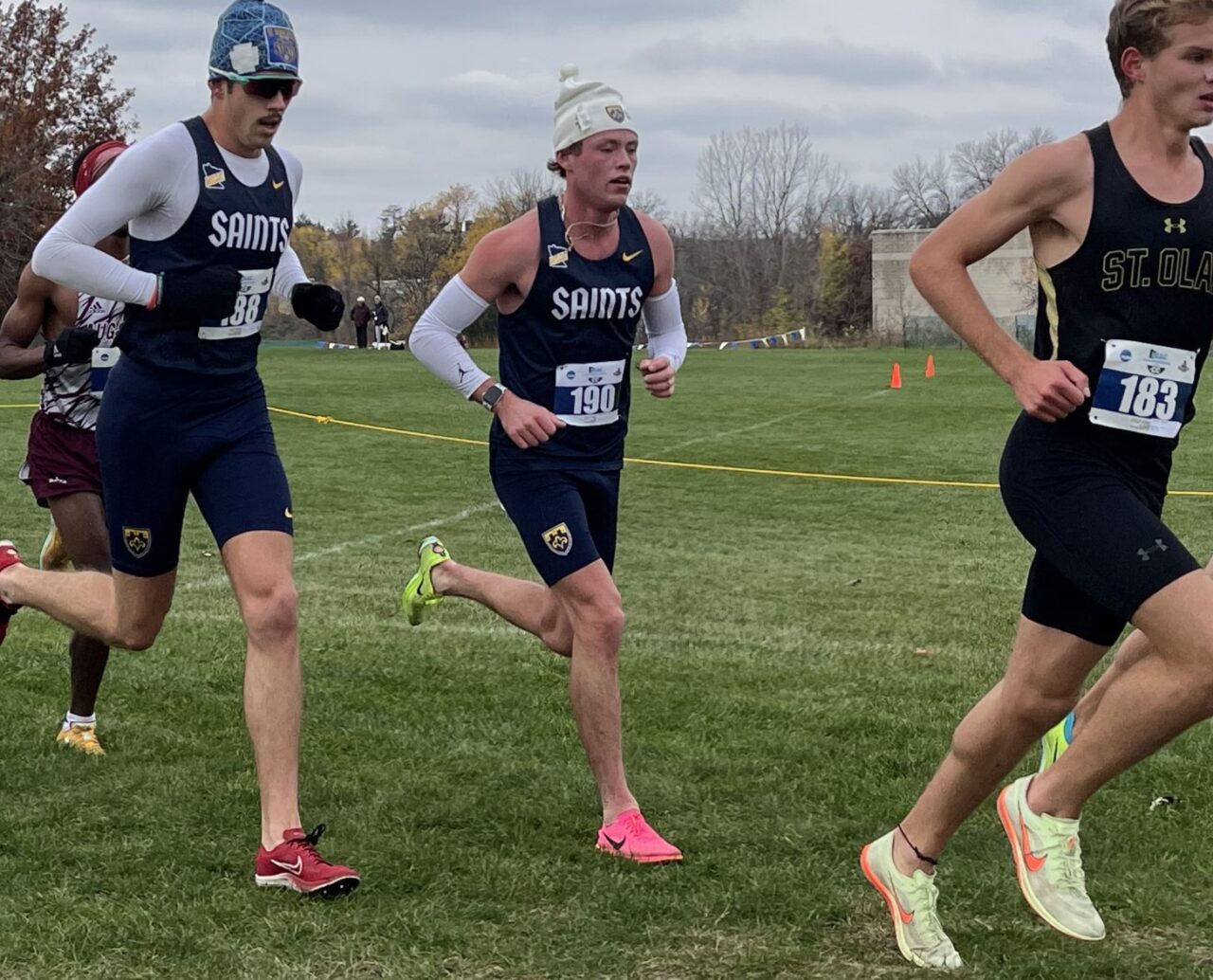 Three college runners on a grass course, the one in the middle wearing a white knit hat, a St. Scholastica singlet and bib 190.