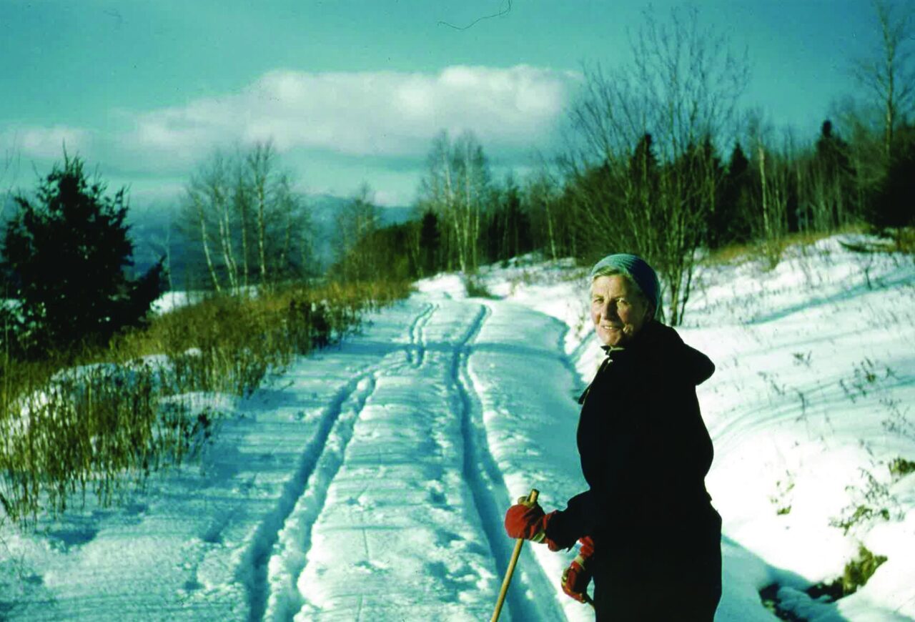 Color archival photo of Maria von Trapp in a dark coat and headscarf holding ski poles beside a snowy ski track
