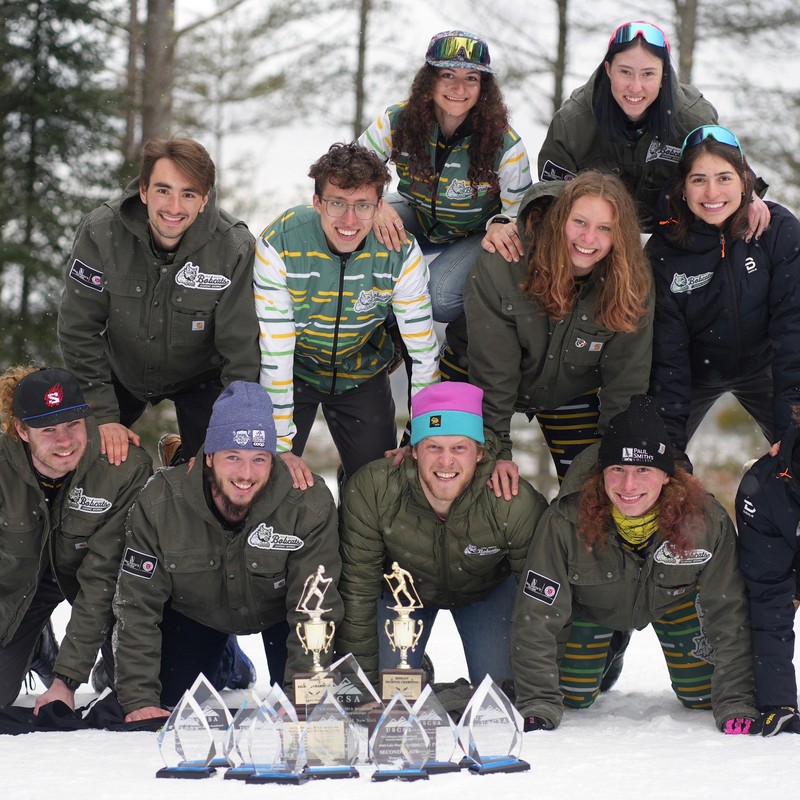 Members of the Paul Smith's College nordic team piled together on snow behind four glass trophies.