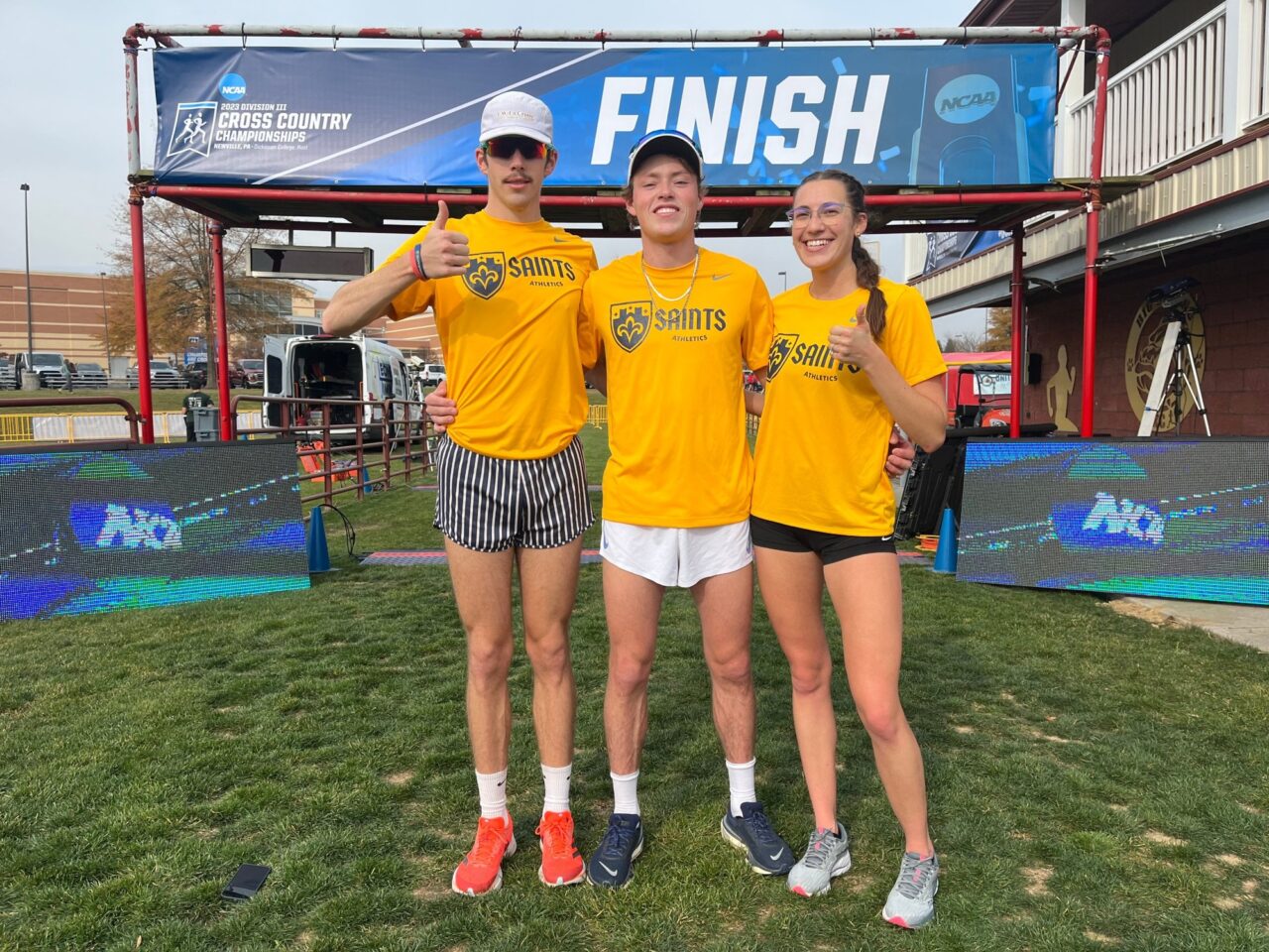 Three St. Scholastica runners in gold Saints shirts smiling and giving a thumbs up beneath an NCAA cross country championships finish banner.