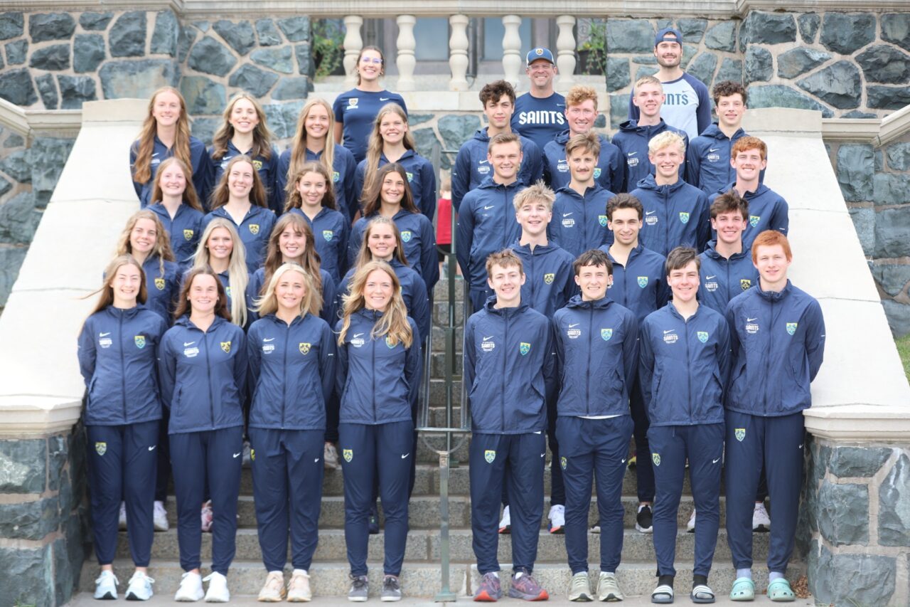 The St. Scholastica cross country team in navy warm-ups posed in rows on stone steps, women on the left and men on the right, with the coaches standing at the top.