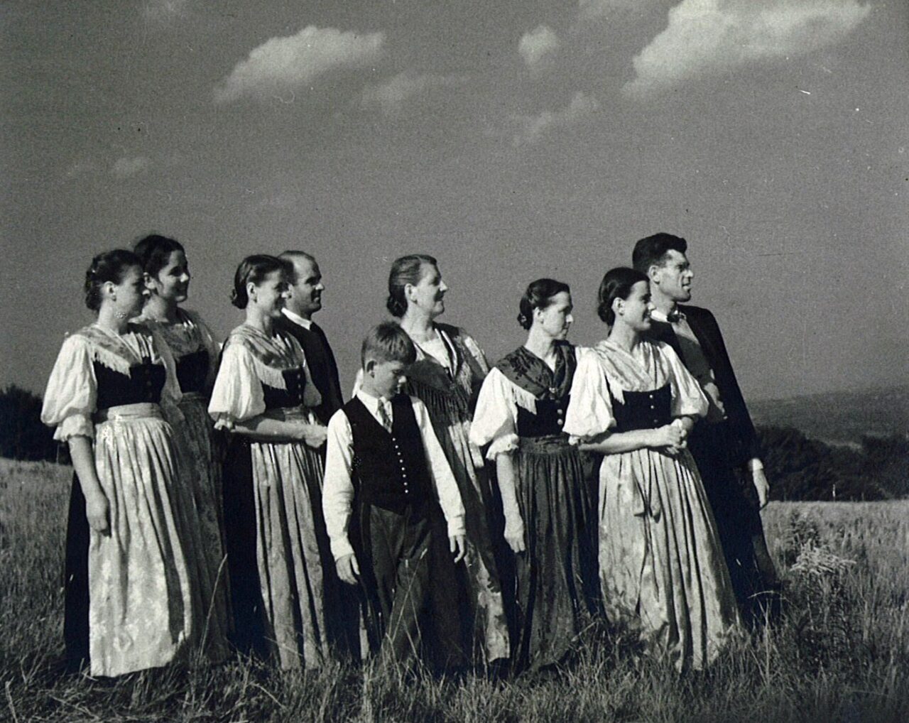 Black and white photo of the Trapp Family Singers standing together in a hillside field, the women in dirndls