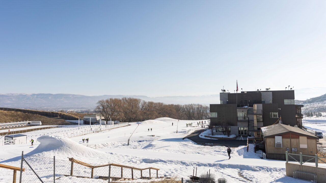 Skiers on snow below the Sports Discovery Center building at Soldier Hollow, with the biathlon range and the Wasatch foothills behind