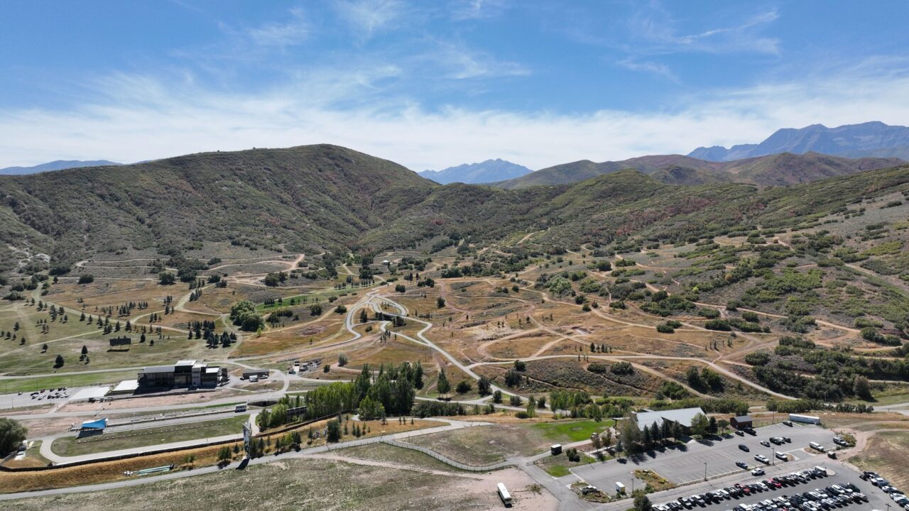 Aerial view of the Soldier Hollow trail network in summer, with lodge buildings, parking and the Wasatch range beyond