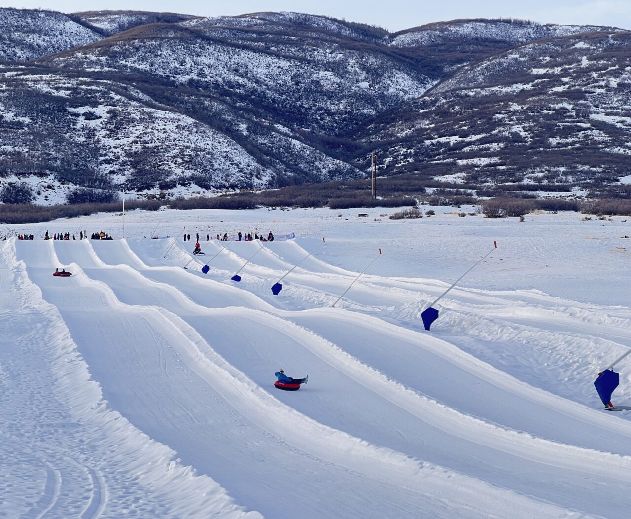 Rows of groomed tubing lanes running down a snow slope at Soldier Hollow with tubers descending and hills behind