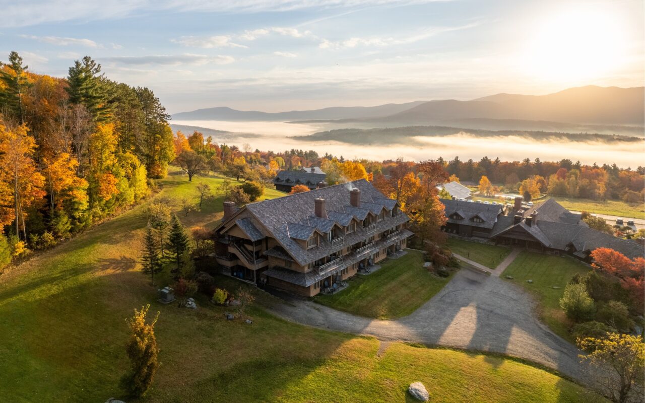 Aerial view of the von Trapp Family Lodge at sunrise, surrounded by fall foliage, with fog in the valley and mountains beyond