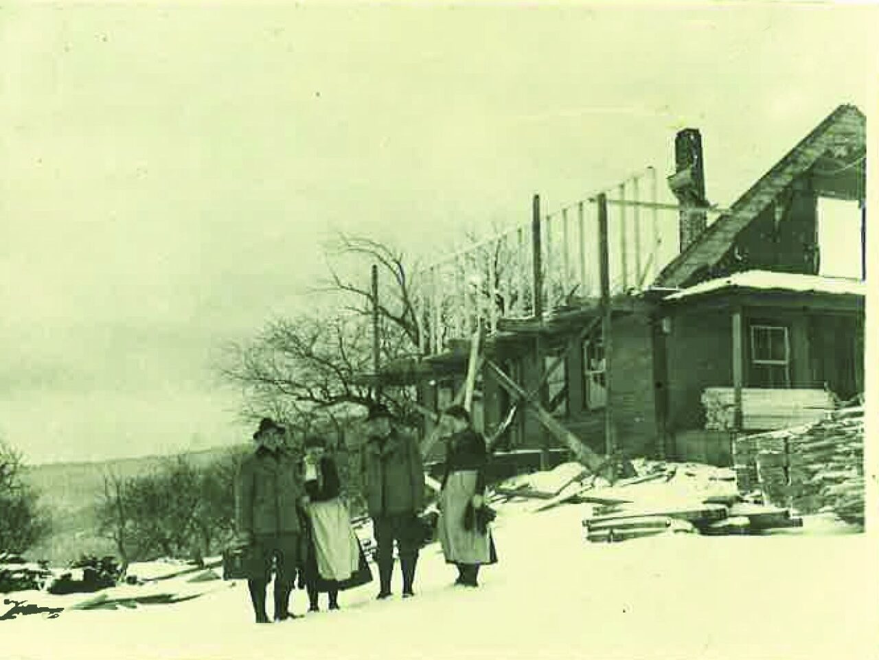 Archival photo of four people standing in the snow in front of a partly built wooden lodge